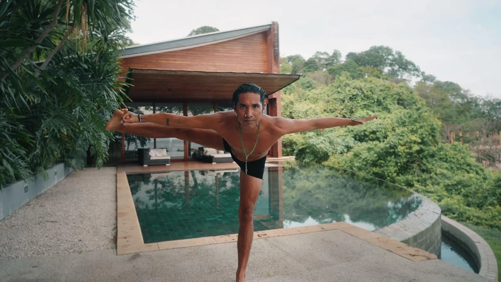 Man balances in a yoga pose on one leg beside an infinity pool with a wooden villa and jungle backdrop