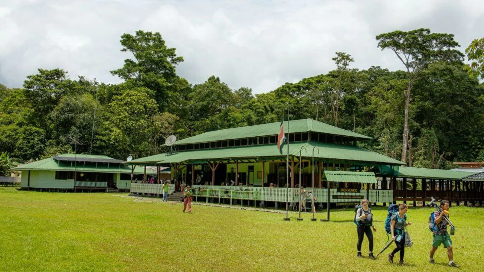 Hikers with backpacks walk across a grassy clearing near a green-roofed ranger station at Sirena Station, Corcovado
