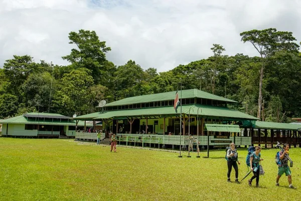 Hikers with backpacks walk across a grassy clearing near a green-roofed ranger station at Sirena Station, Corcovado