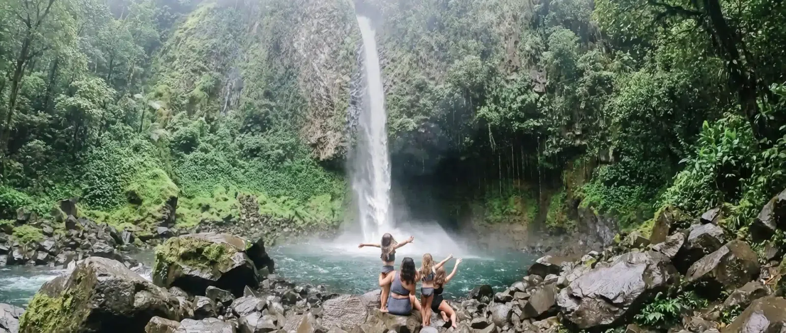 Three women sit on rocks admiring a tall waterfall flowing into a jungle pool in Costa Rica