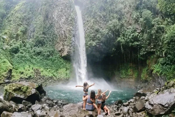 Three women sit on rocks admiring a tall waterfall flowing into a jungle pool in Costa Rica
