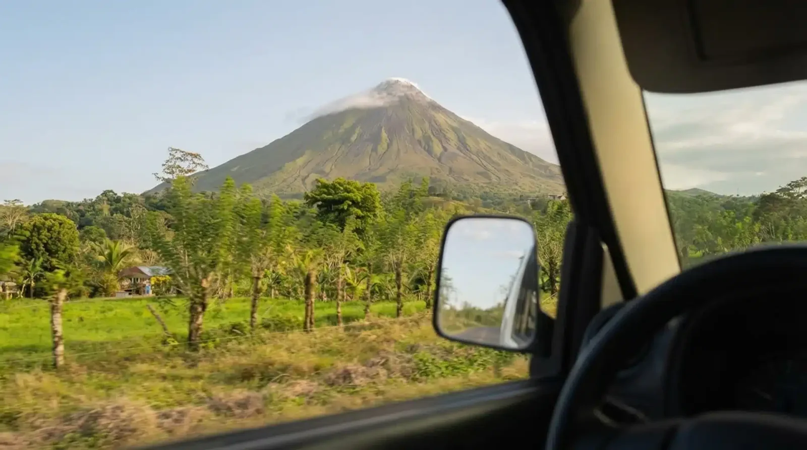 Arenal Volcano seen through a car window and side mirror, with green fields and trees in the foreground