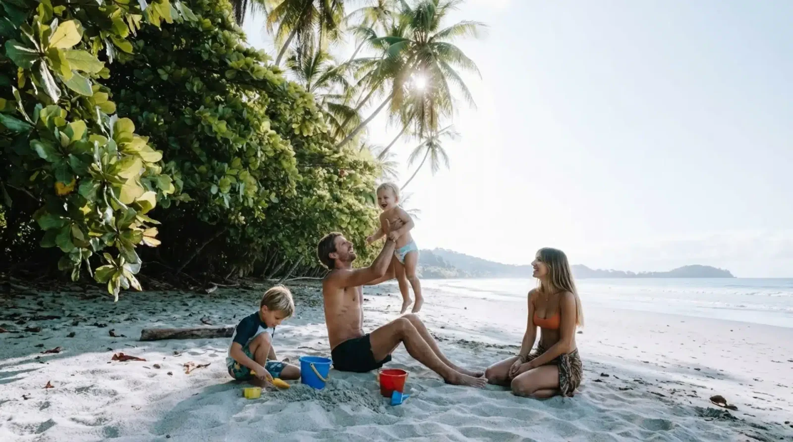 Family plays on a palm-fringed beach, with a father lifting a toddler as a boy digs in the sand