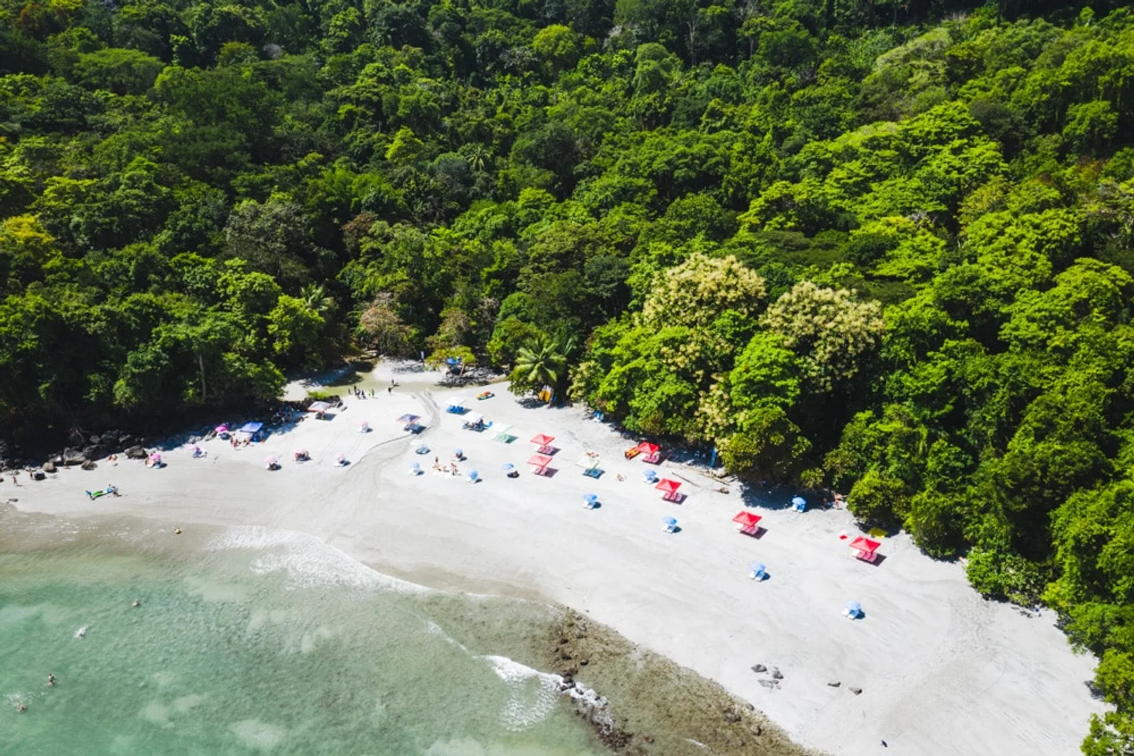 Aerial view of a white sand beach with colorful umbrellas, fringed by dense green rainforest near Manuel Antonio, Costa Rica