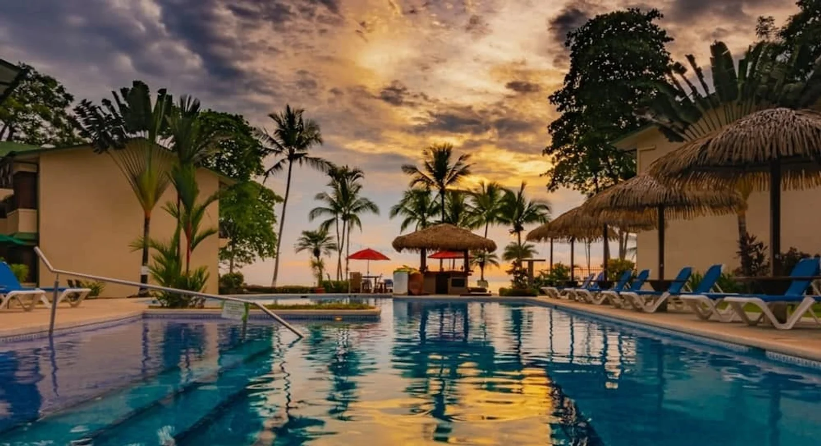 Resort pool with palm trees and a thatched-roof bar at sunset in Jacó