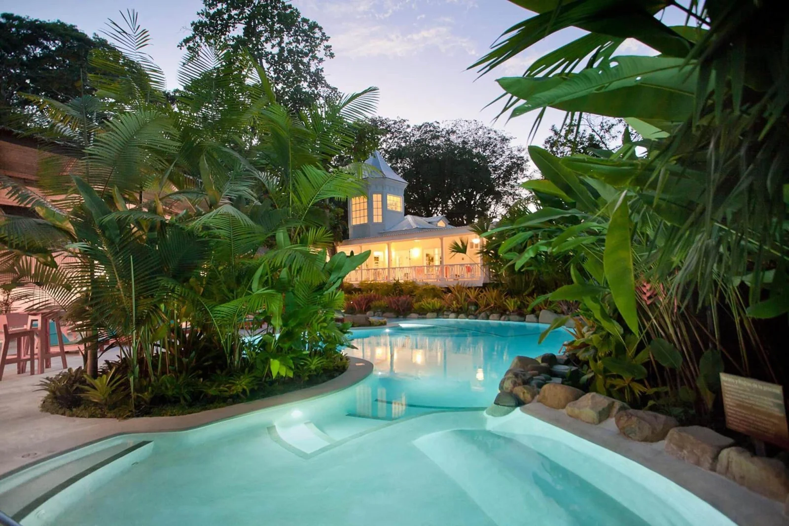 Lit pool winds through lush tropical gardens with a white gazebo-style building glowing at dusk