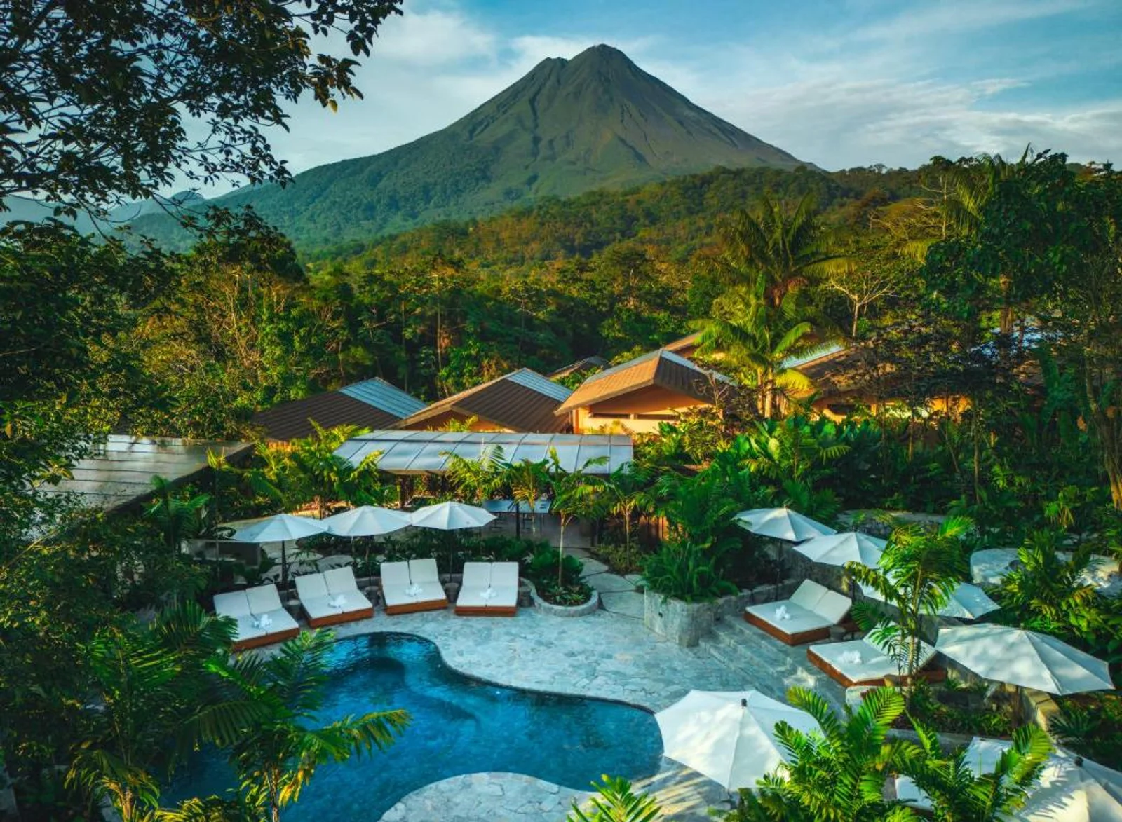 Resort pool with white loungers and umbrellas surrounded by jungle, with Arenal Volcano rising behind
