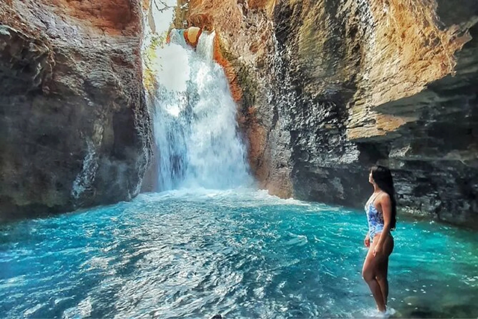 Woman standing in a turquoise pool inside a rocky canyon, facing a tall waterfall