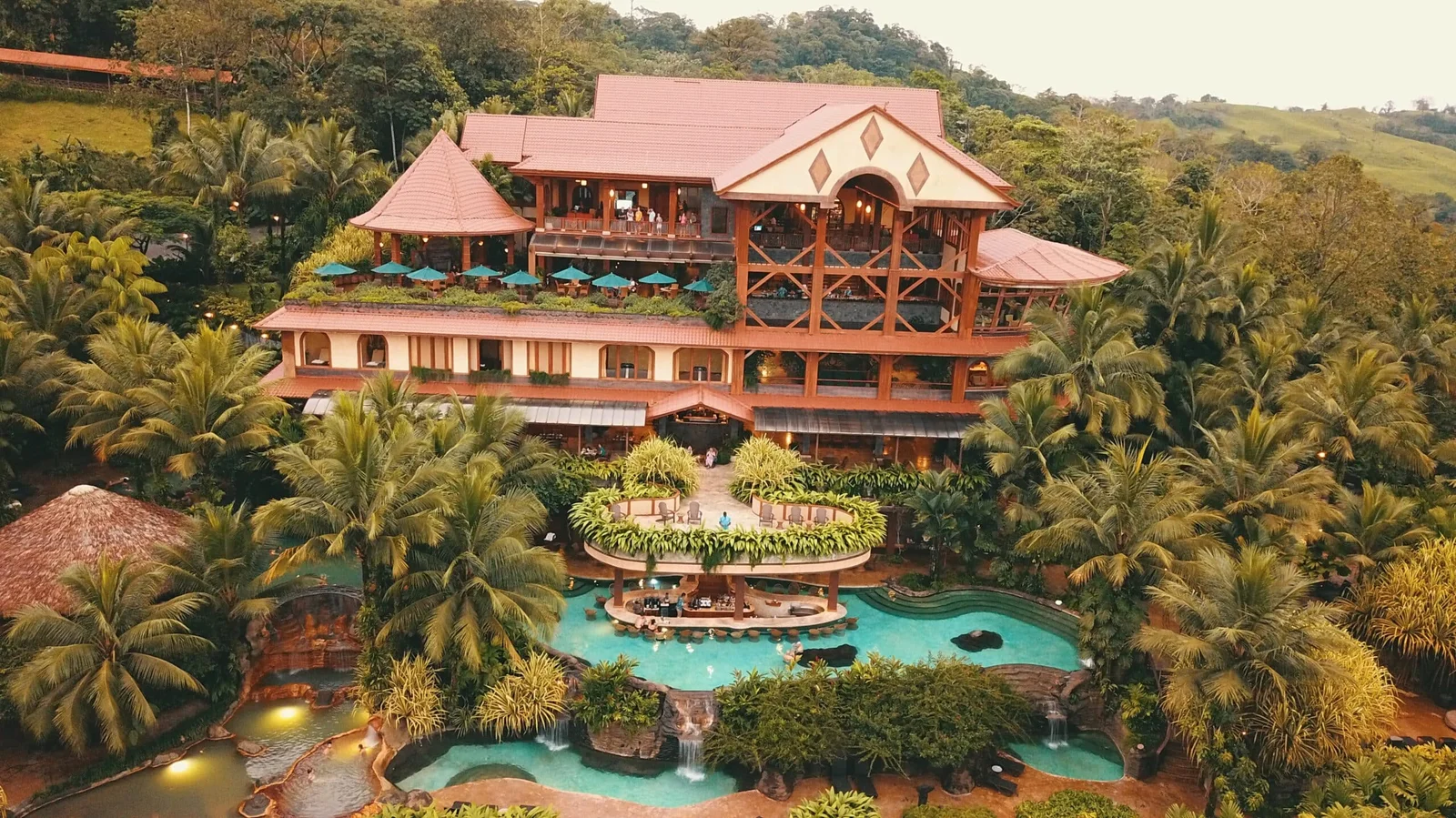 Aerial view of a jungle lodge with red rooftops, pools, and waterfalls surrounded by rainforest