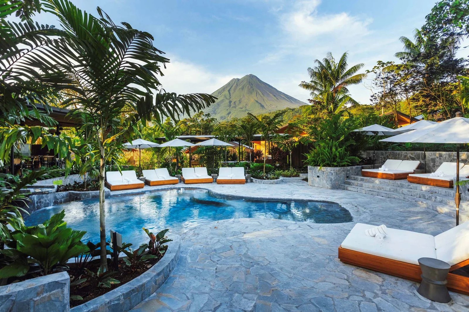 Resort pool with lounge chairs and umbrellas, tropical plants, and Arenal Volcano in the background