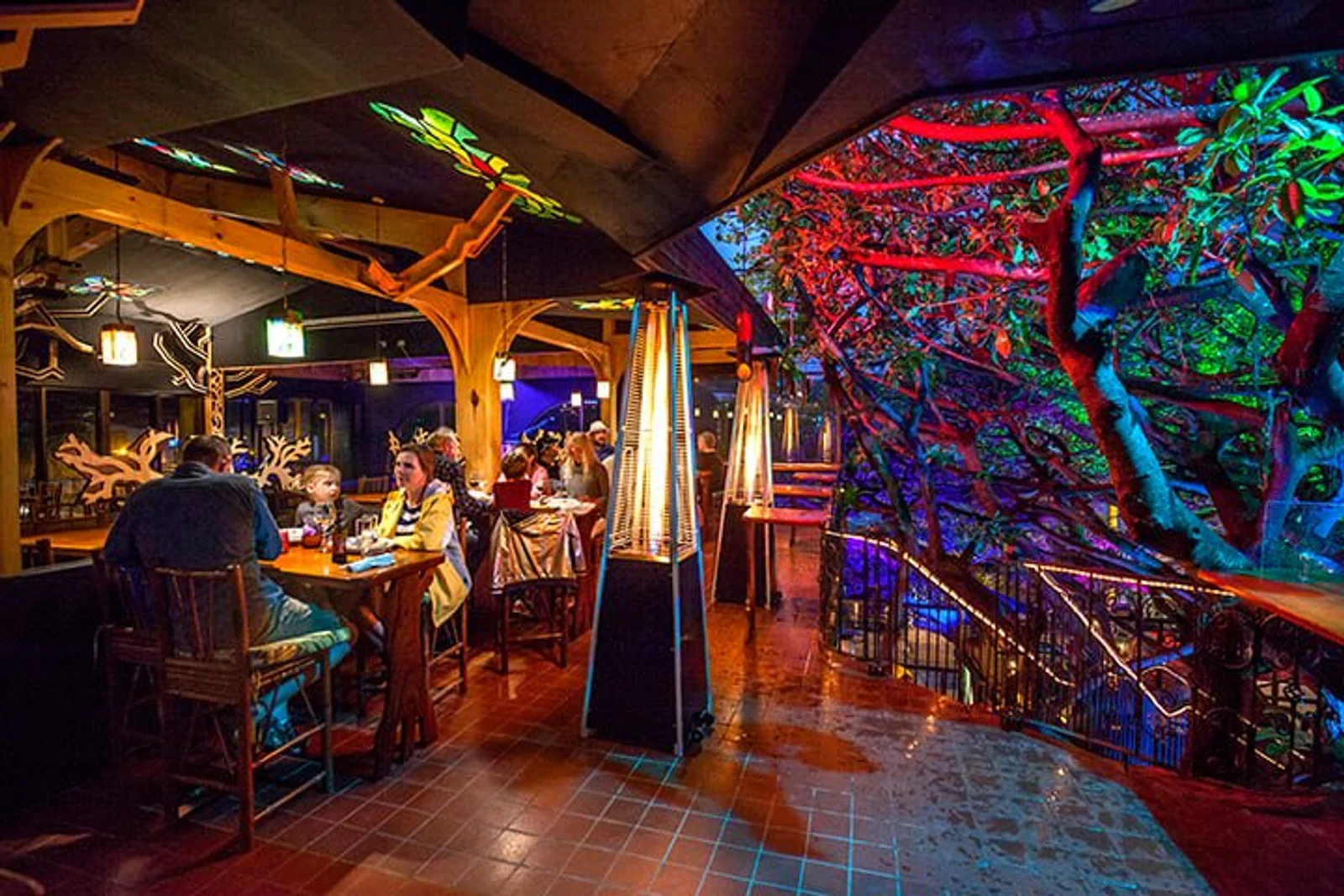 Open-air restaurant at night with wooden tables, patio heaters, and colorful lights illuminating trees over a railed terrace