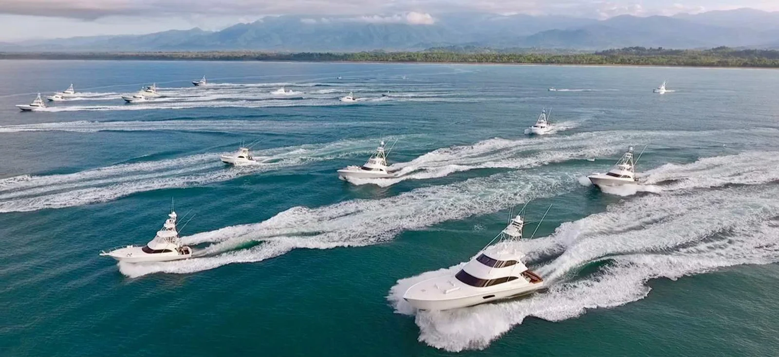 Fleet of sport fishing yachts racing across the ocean with wakes trailing, coastline and mountains behind