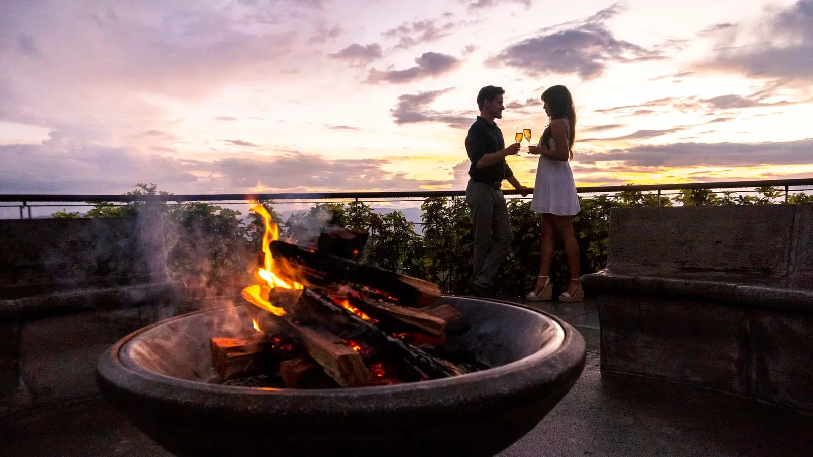 Couple toasting wine glasses by a rooftop fire pit at sunset