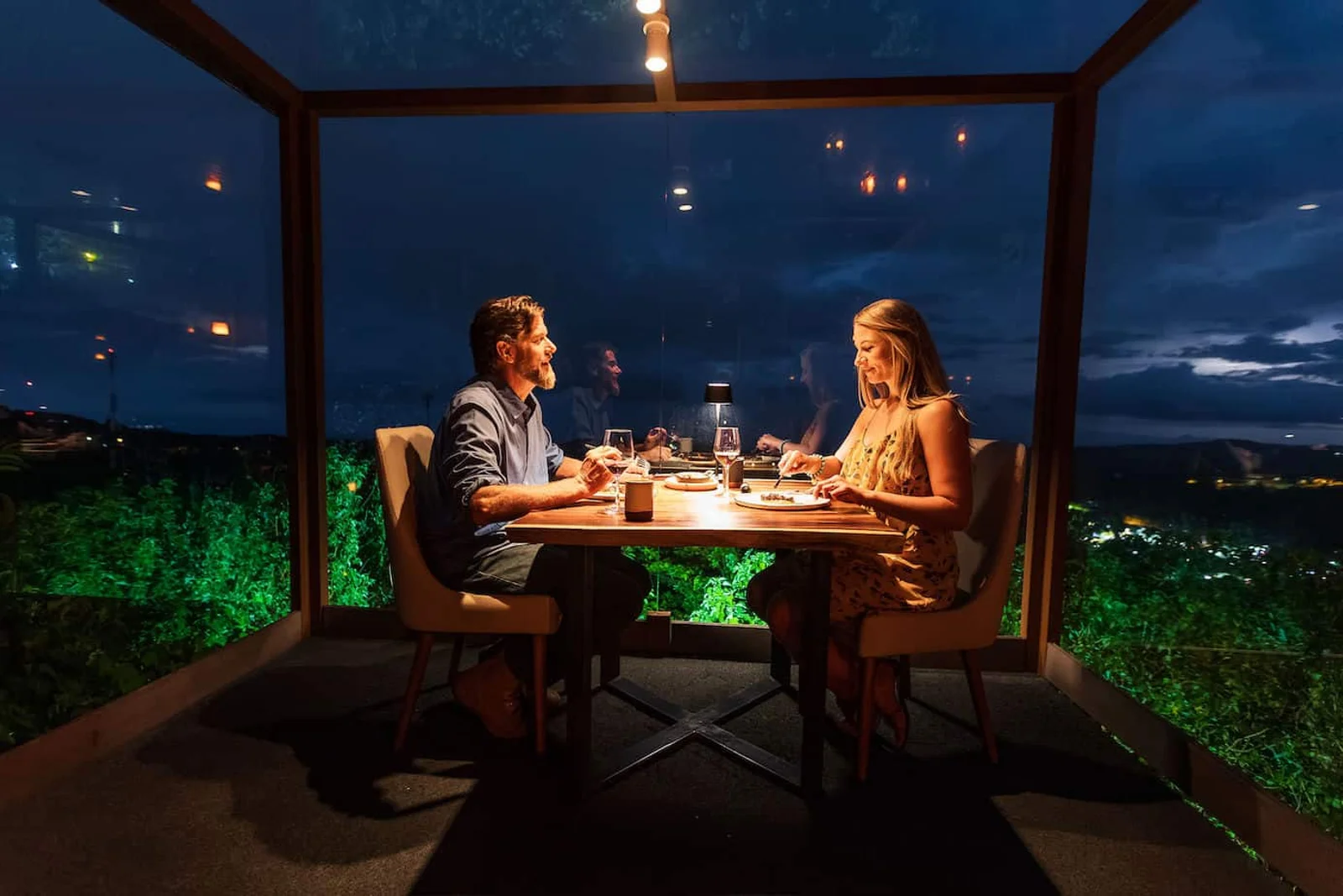 Couple dining at a glass-walled table overlooking city lights and hills at night