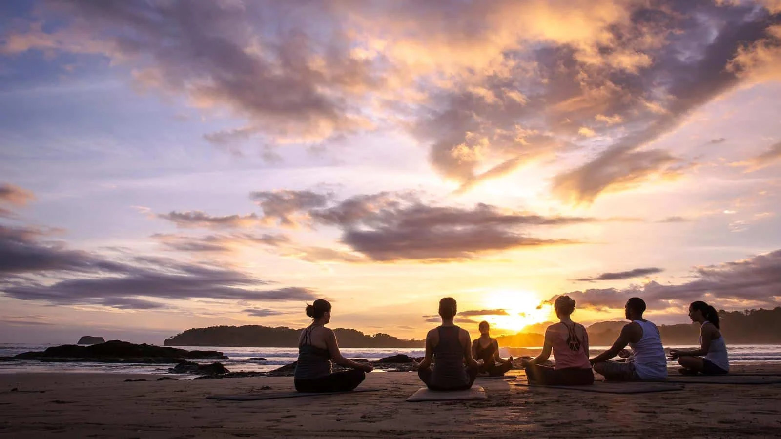 Group sits in meditation poses on yoga mats on a beach, silhouetted against a colorful sunset