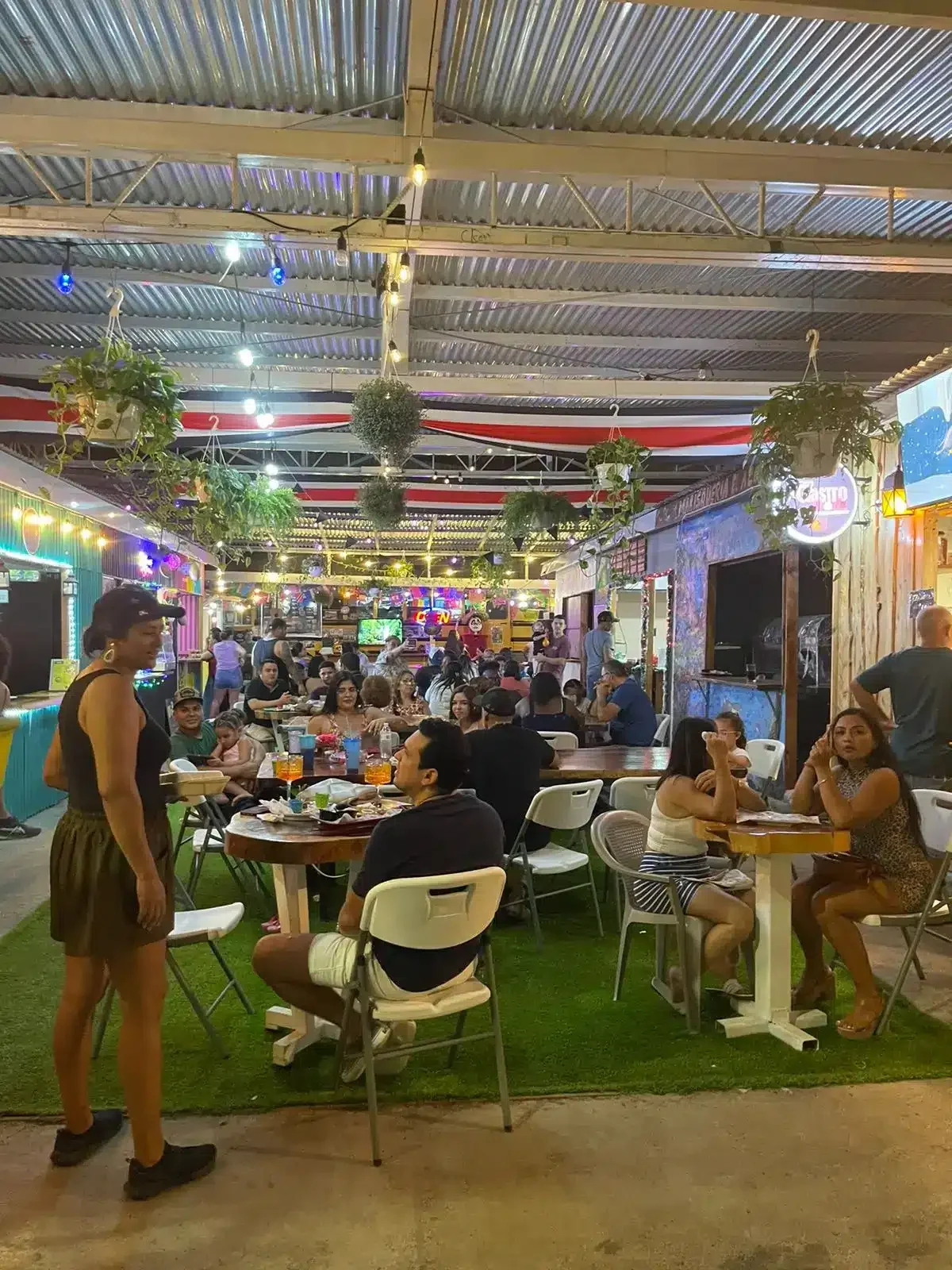 Diners sit at tables on artificial turf inside an open-air food court with hanging plants and a corrugated metal roof