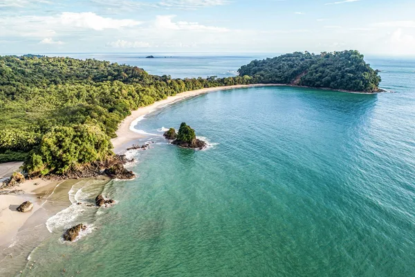 Aerial view of a curved sandy beach, turquoise cove and forested headlands in Manuel Antonio