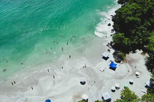 Aerial view of a tropical beach cove with turquoise water, swimmers, sun umbrellas and forested cliffs