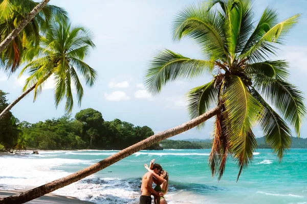 Couple embracing on a tropical beach beneath leaning palm trees with turquoise water