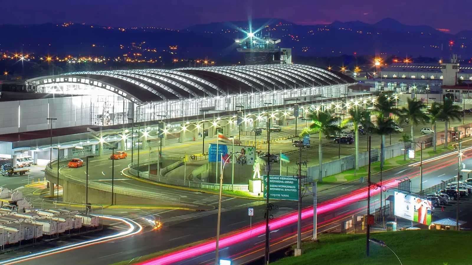 Airport terminal at night with curved illuminated roof, palm trees, and light trails from traffic on the entrance road