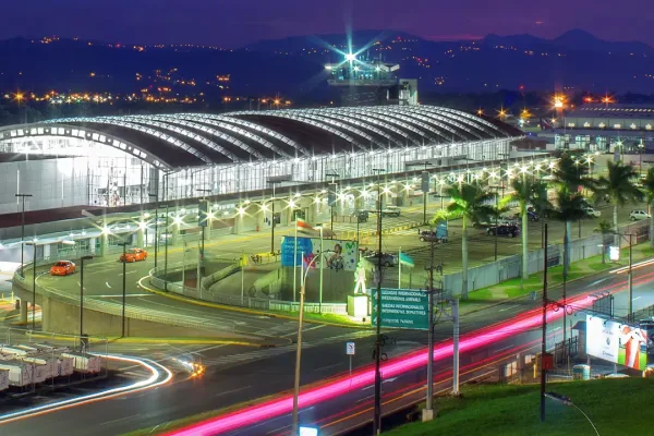 Airport terminal at night with curved illuminated roof, palm trees, and light trails from traffic on the entrance road