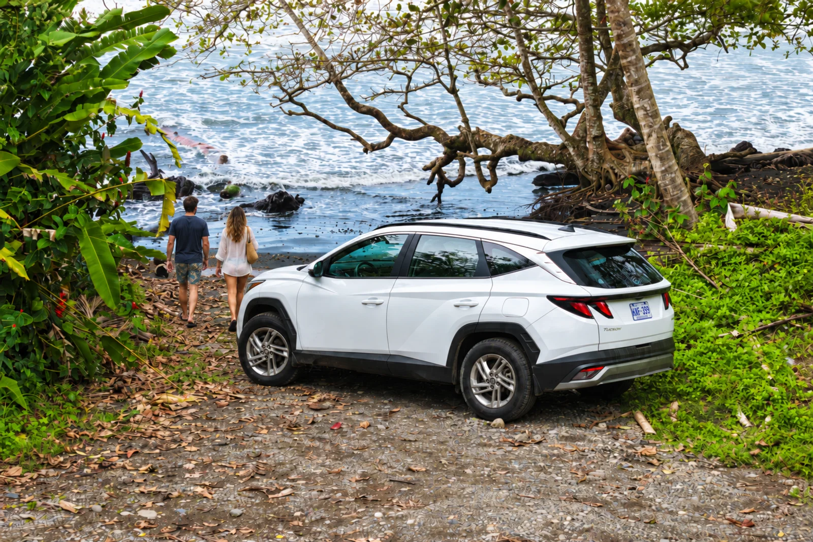 White SUV parked on a gravel path near a beach, with two people walking toward the shoreline