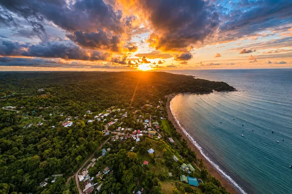 Aerial view of a coastal village nestled in rainforest at sunset, with a curved beach and calm ocean