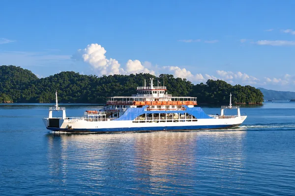 A car ferry crosses calm blue water with forested islands and hills in the background