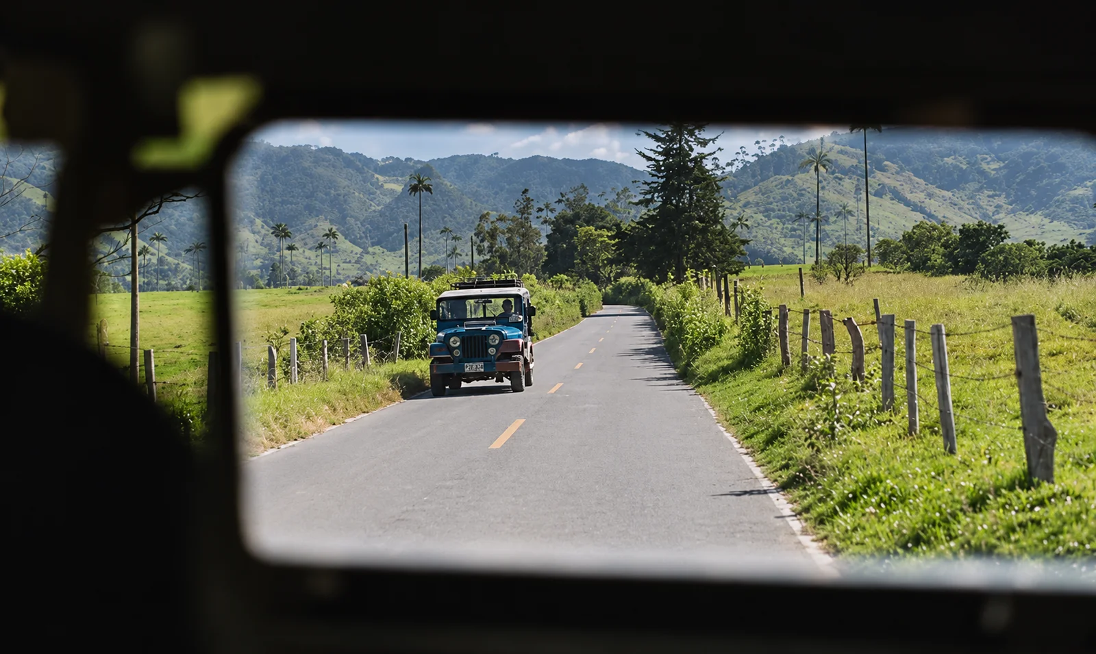 A blue jeep on a rural road seen through a vehicle window, with palm-dotted hills in the distance