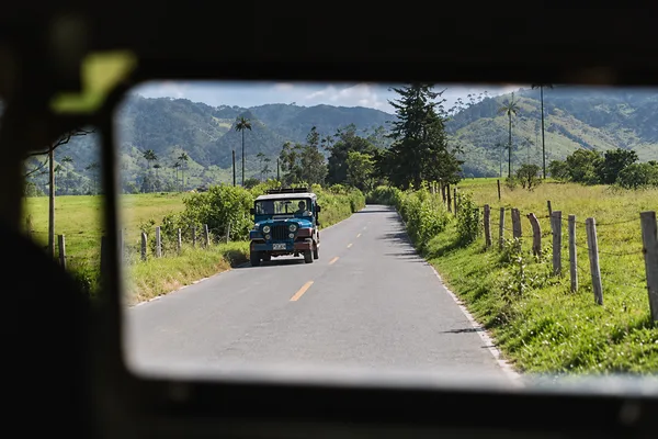A blue jeep on a rural road seen through a vehicle window, with palm-dotted hills in the distance