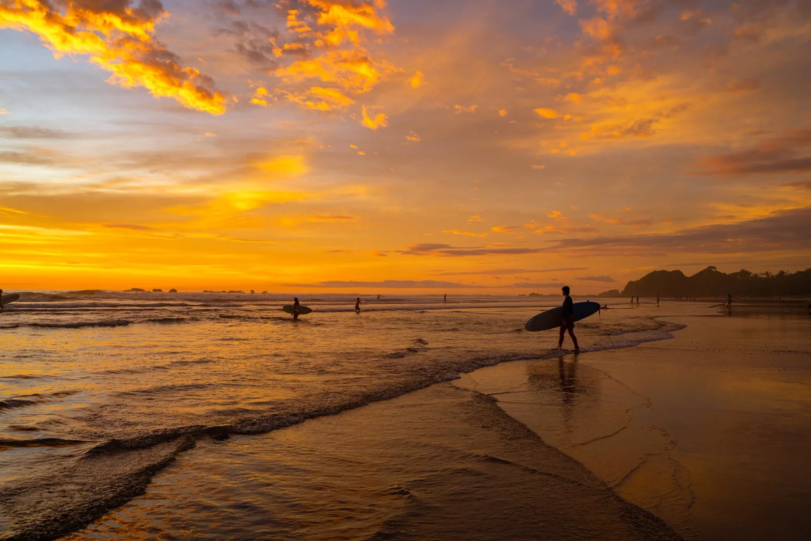 Surfer carrying a board walks along the shoreline as a vivid orange sunset lights up the sky over the beach
