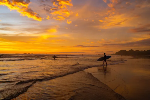 Surfer carrying a board walks along the shoreline as a vivid orange sunset lights up the sky over the beach