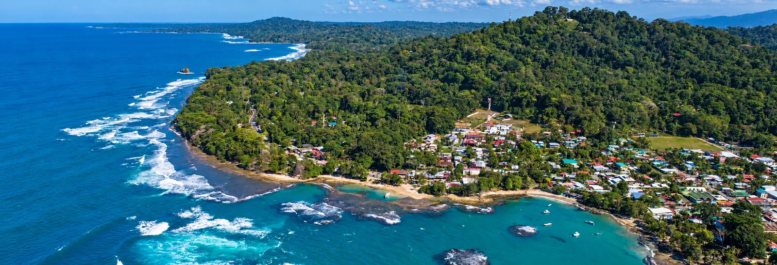 Aerial view of a coastal village nestled between rainforest hills and a turquoise bay with coral reefs