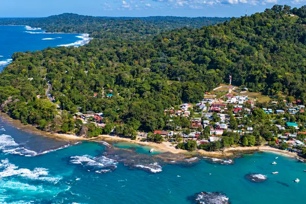 Aerial view of a coastal village nestled between rainforest hills and a turquoise bay with coral reefs