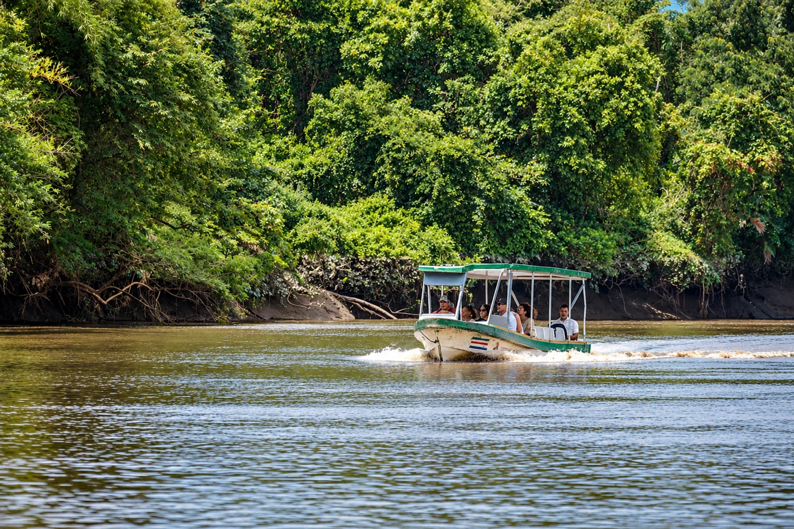 A covered tour boat carrying passengers cruises along a tree-lined river