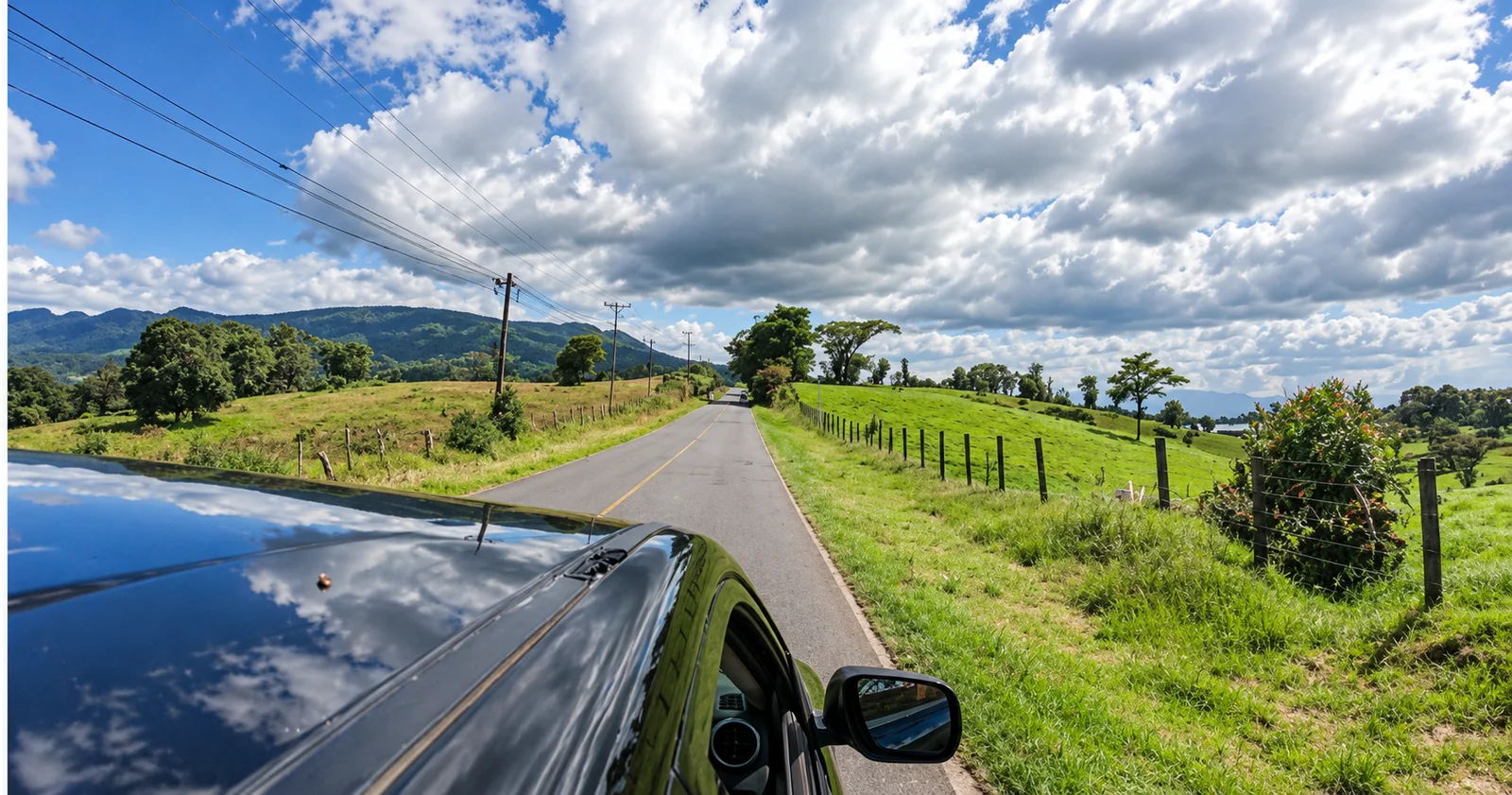 Rural paved road through green countryside with hills, seen from a car with the hood and side mirror visible