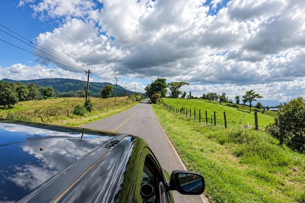 Rural paved road through green countryside with hills, seen from a car with the hood and side mirror visible
