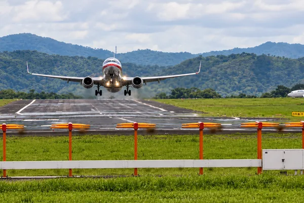 A jet airliner touches down on a runway with forested mountains in the background