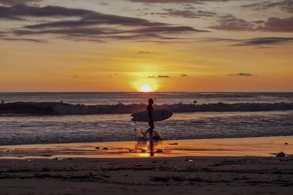 Silhouette of a surfer carrying a surfboard along the beach as the sun sets over the ocean