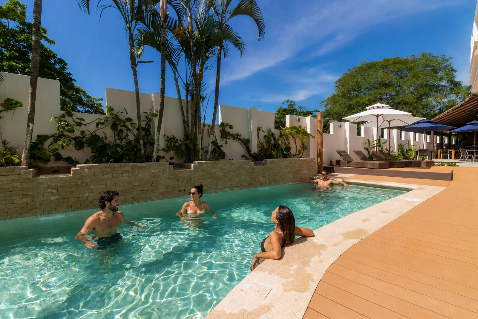 Guests relax in a resort pool lined with palm trees and a stone wall, with wooden deck and umbrellas nearby