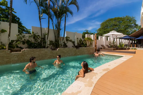 Guests relax in a resort pool lined with palm trees and a stone wall, with wooden deck and umbrellas nearby