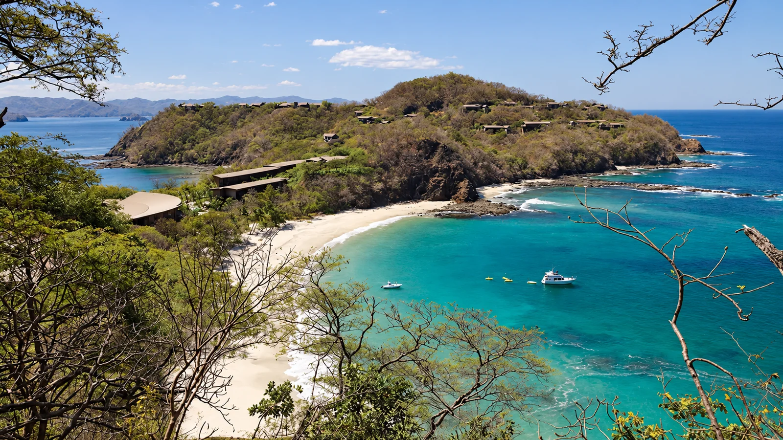 Aerial view of a turquoise bay with a white sand beach, forested headland, and boats anchored offshore