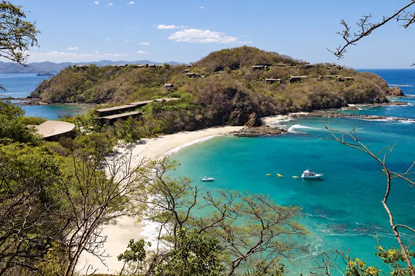 Aerial view of a turquoise bay with a white sand beach, forested headland, and boats anchored offshore