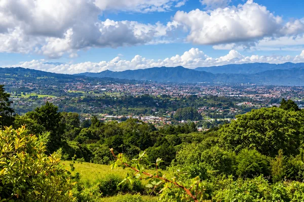 Sprawling valley town seen from a green hillside, with mountains and clouds in the distance near San Jose