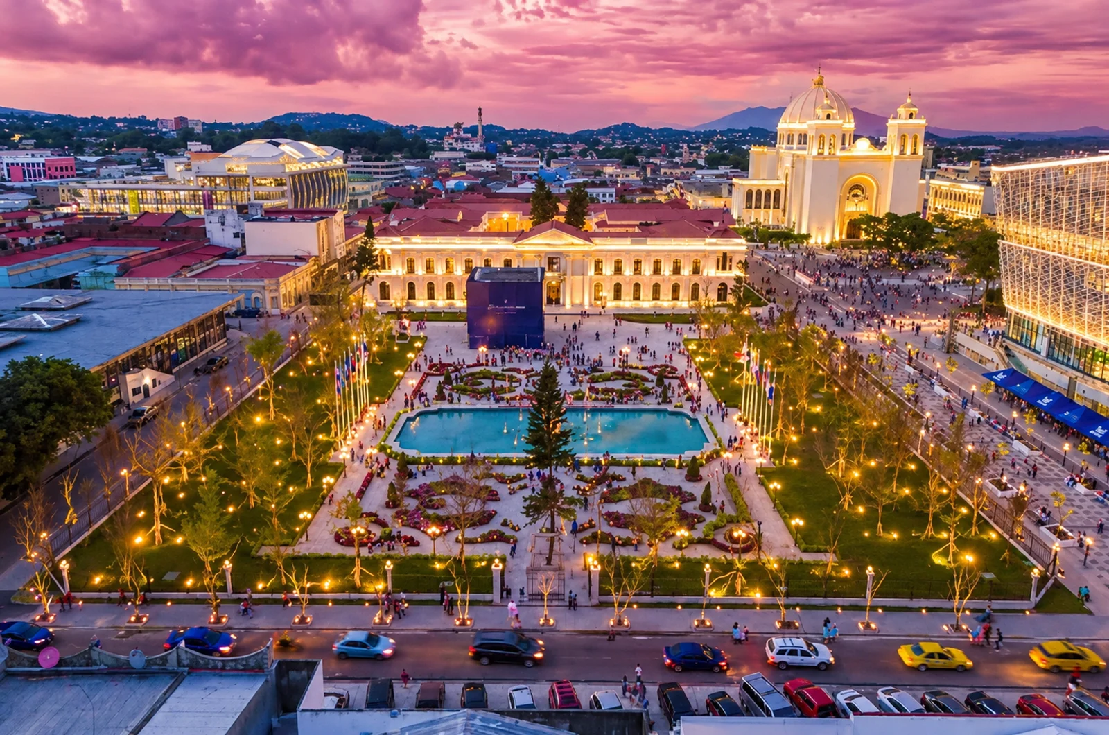 Aerial twilight view of San Salvador's central plaza with illuminated gardens, pool, cathedral and city skyline