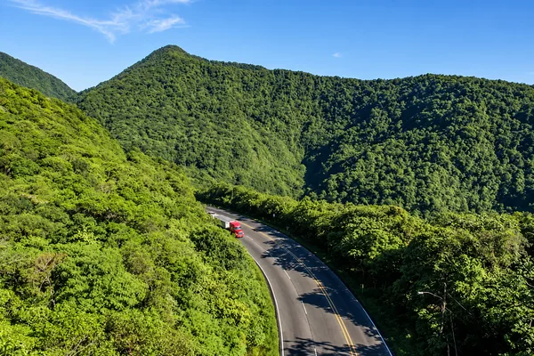 A truck drives along a winding mountain road through dense green forested hills
