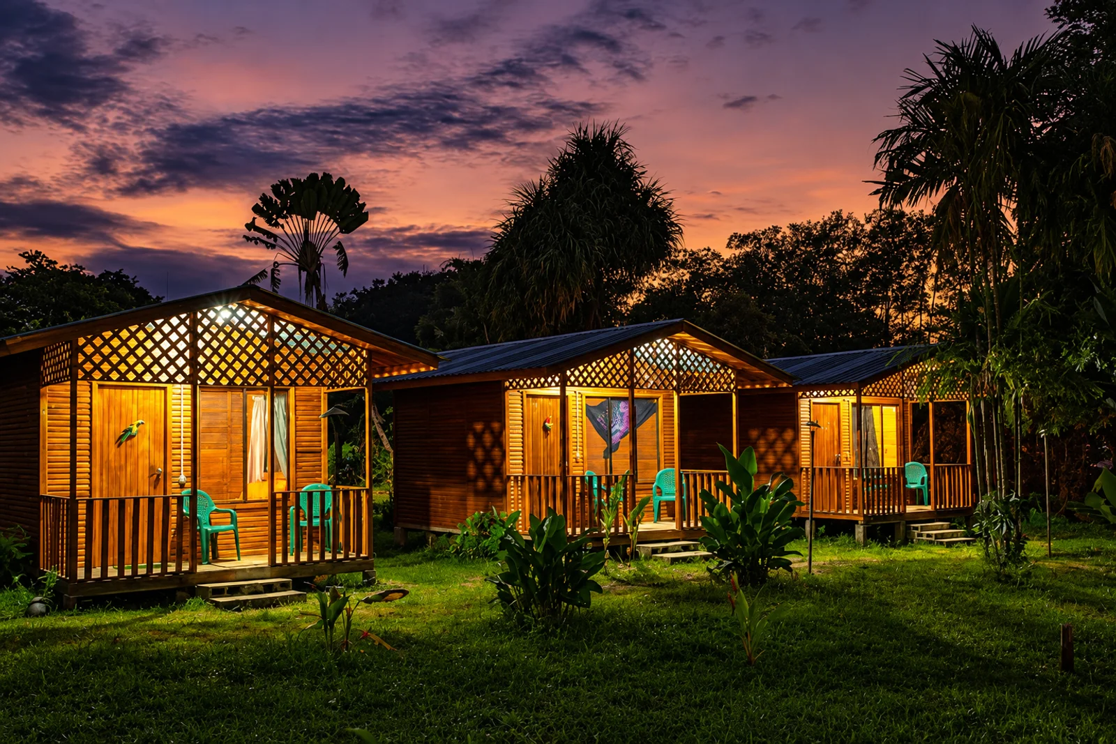 Wooden cabins with lattice porches glow at dusk under a pink and purple sky, surrounded by palm trees