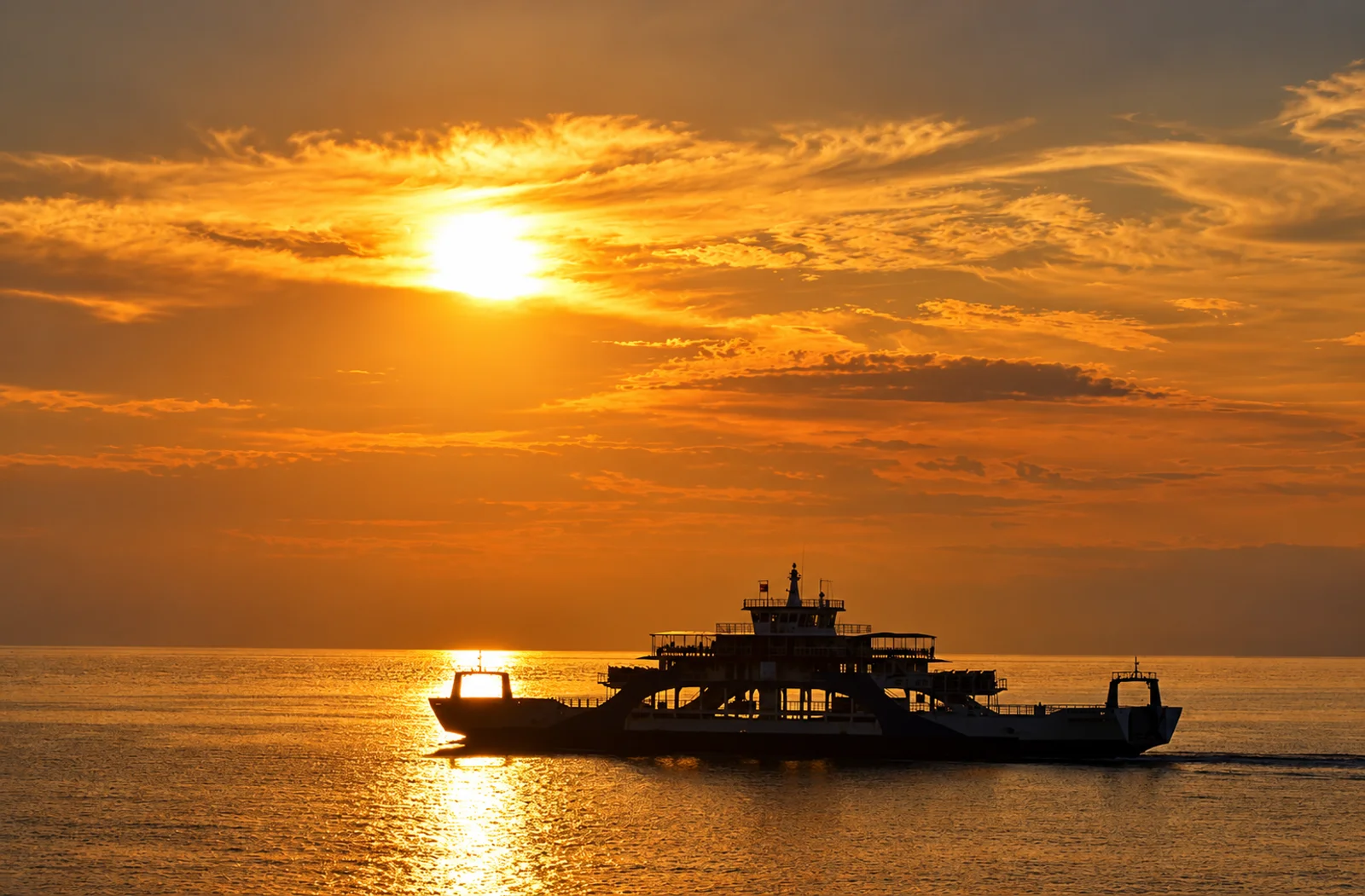 Silhouette of a car ferry crossing the water at sunset with a glowing orange sky