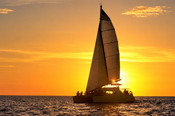 Silhouette of a sailing catamaran with raised sail against an orange sunset over the ocean