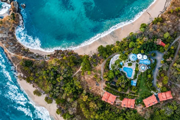 Aerial view of a beachfront resort with a round pool, domed buildings, and red-roofed villas amid trees by the ocean
